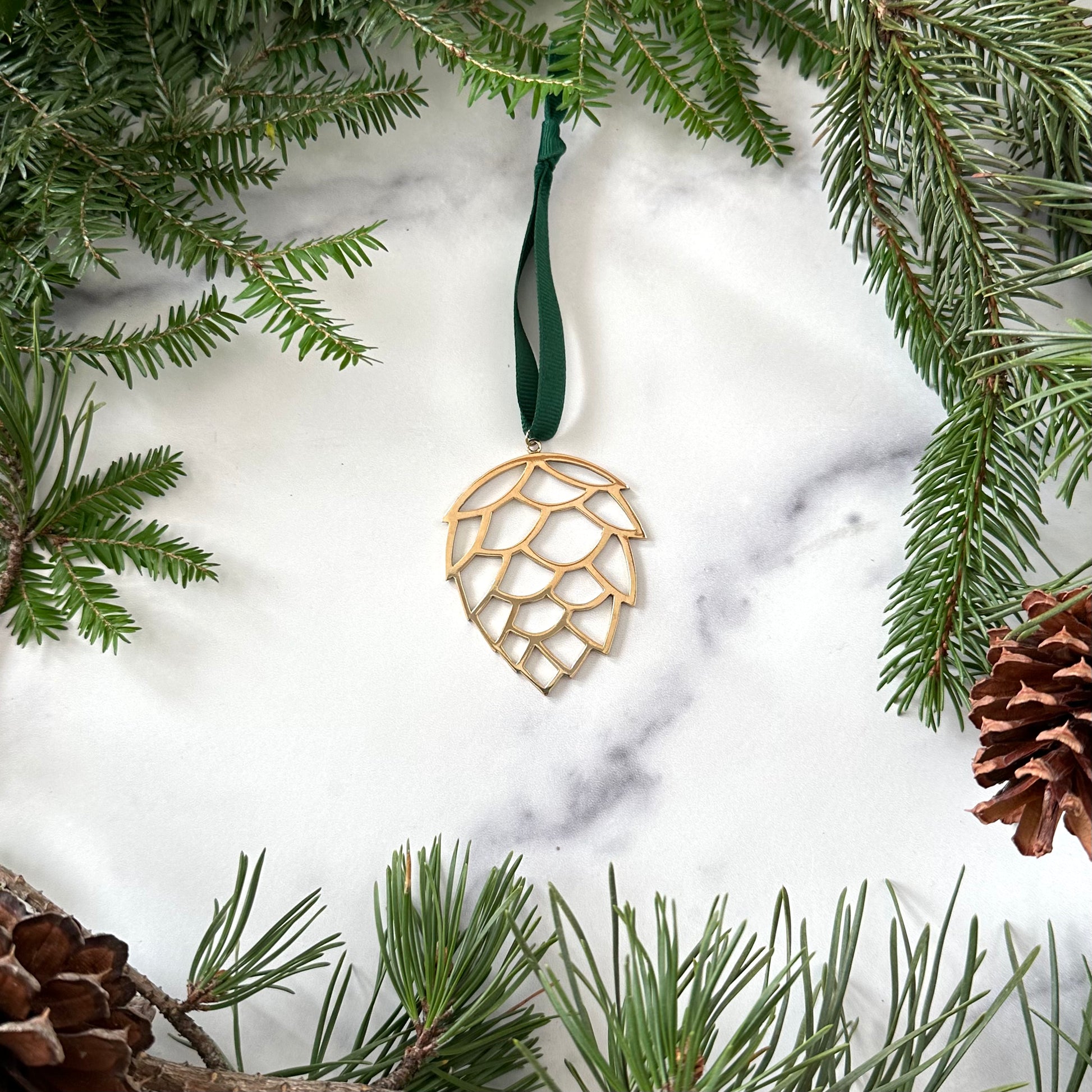 A handmade brass pinecone ornament on a marble background surrounded by greenery and pinecones