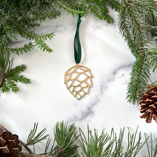 A handmade brass pinecone ornament on a marble background surrounded by greenery and pinecones