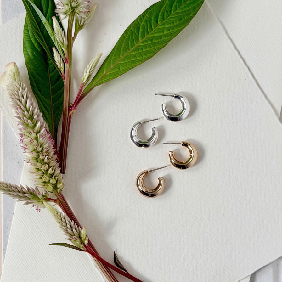 Four small hoop earrings in silver and gold finishes, arranged on a textured white paper background alongside green leaves and pinkish flower stems.