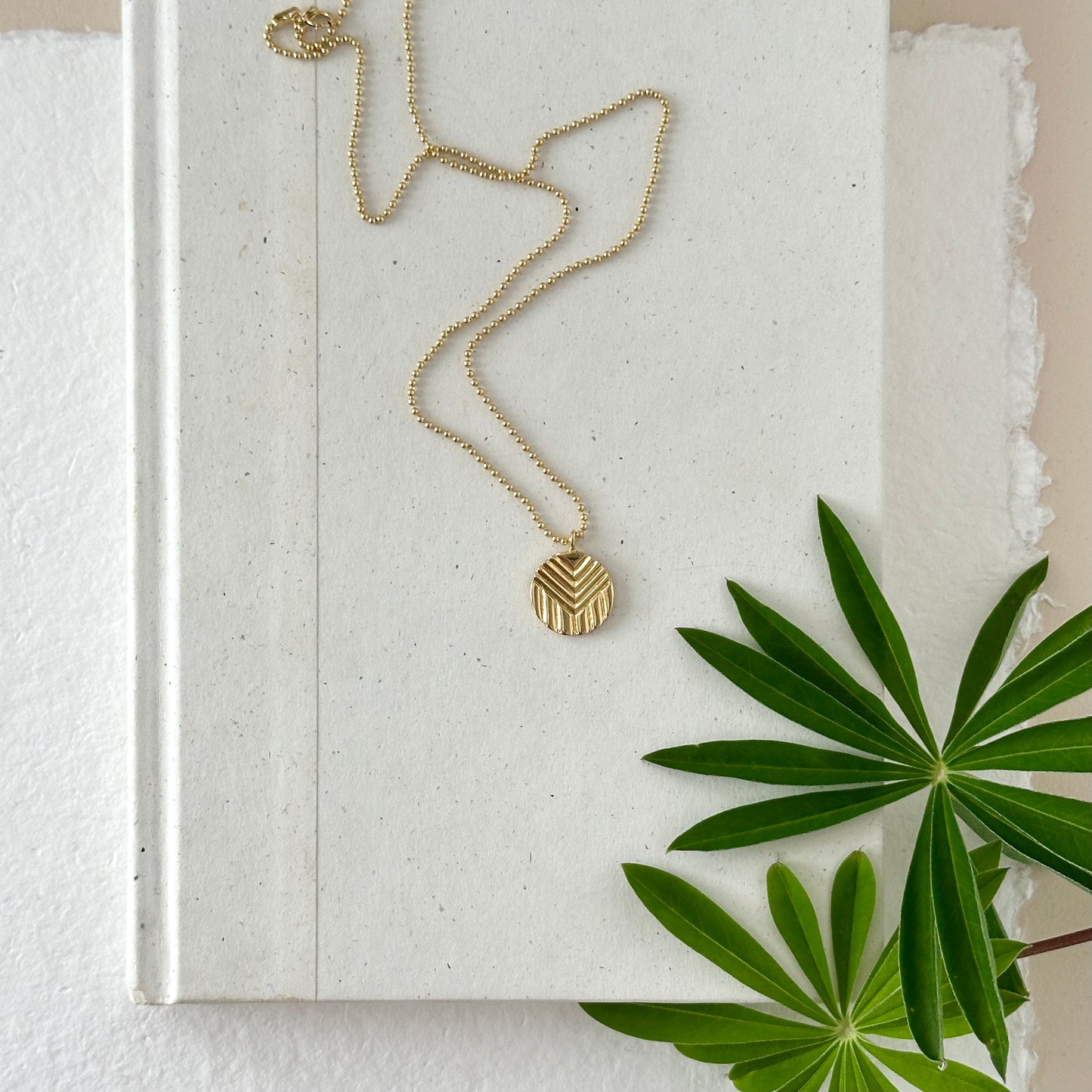 A gold progress pride pendant on a gold ball with chain by Sarah Cecelia. The pendant and chain are on a neutral background with greenery.