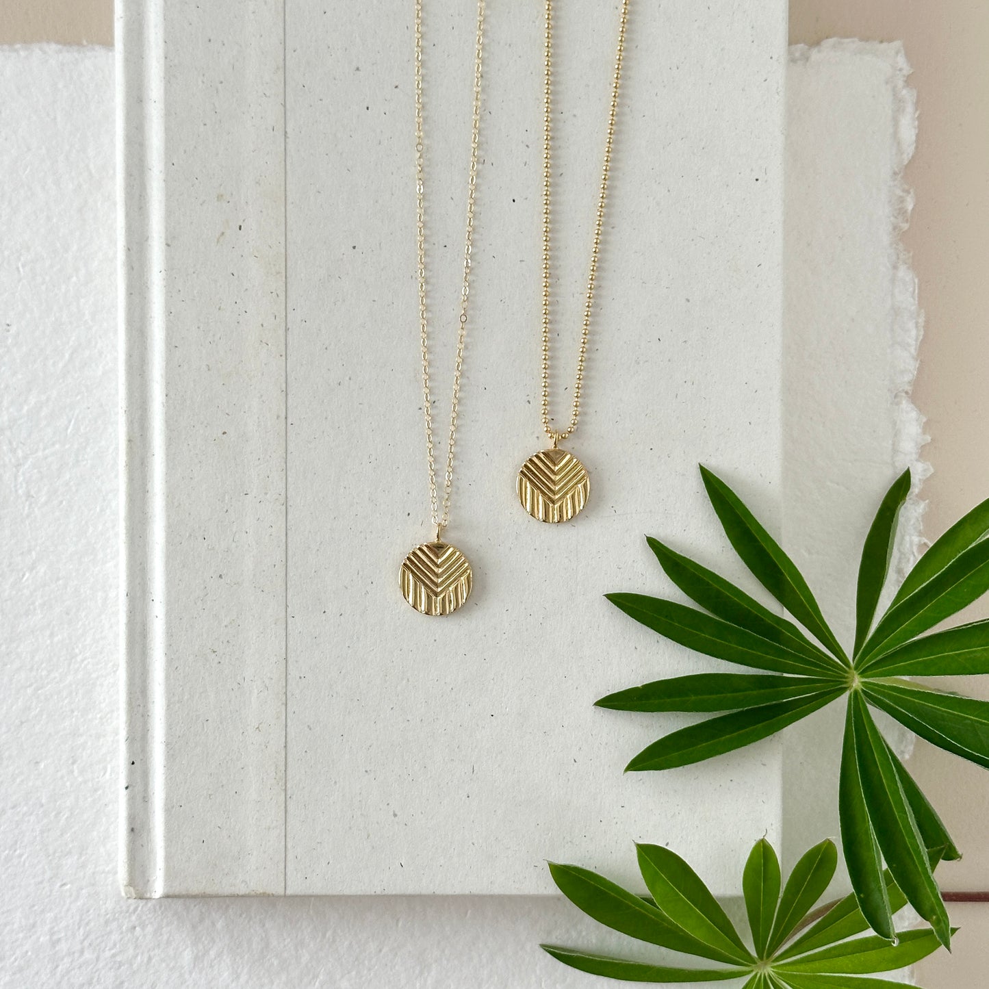Two gold progress pride pendants on chains with neutral background and greenery.
