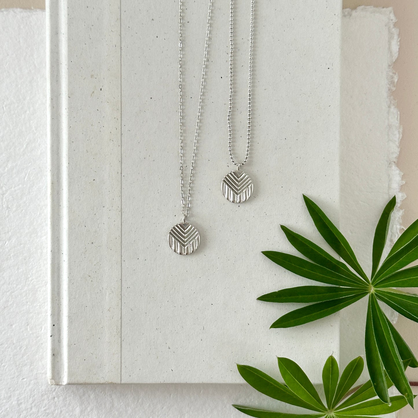 Two sterling silver progress pride flag pendants with silver chains on a neutral background with greenery.