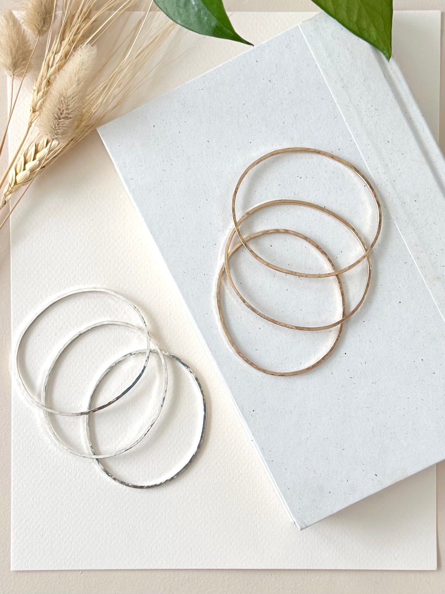 three gold and three silver handmade bangle bracelets displayed on a book and an ivory background with greenery and dried grasses