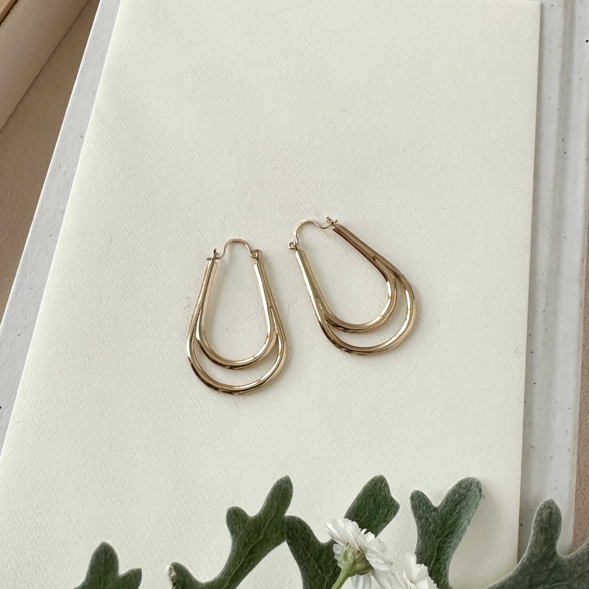 Gold hoop earrings on a white surface with green leaves in the background