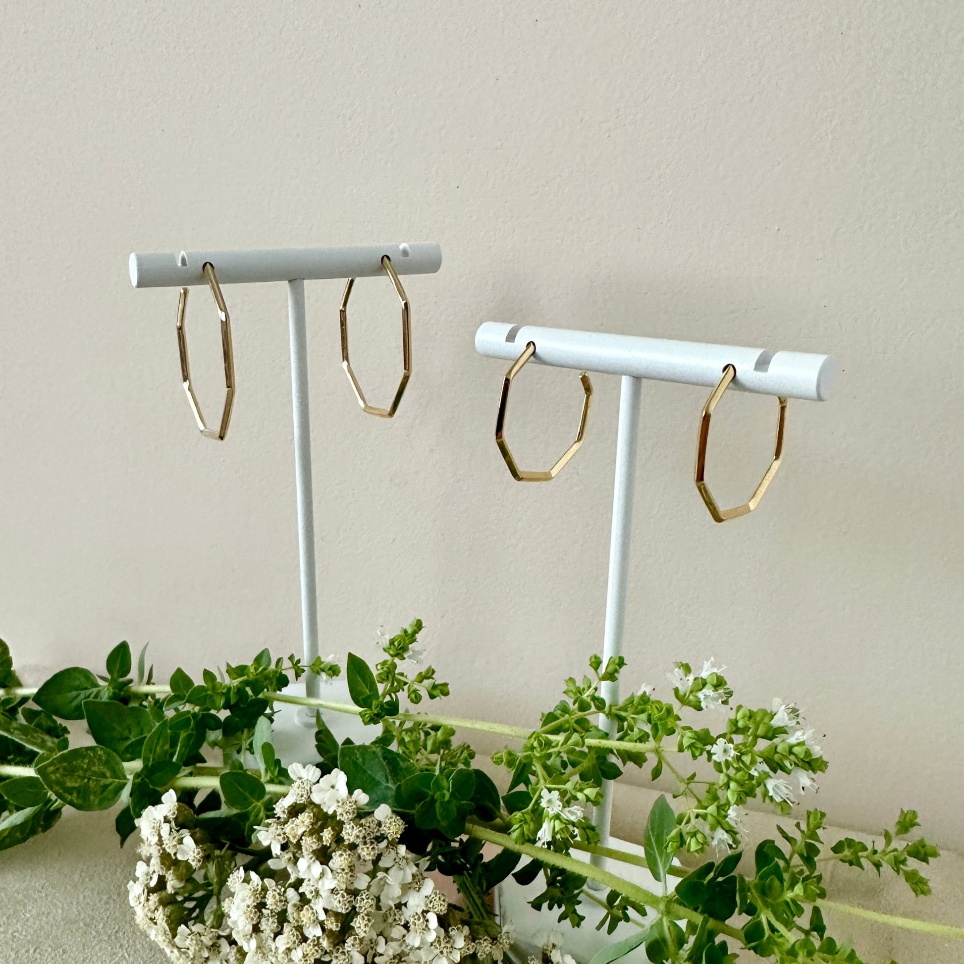 Gold hoop earrings on white stands with a light background and greenery.