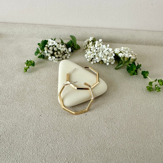 Gold geometric earrings on a white stone with small white flowers and green leaves.