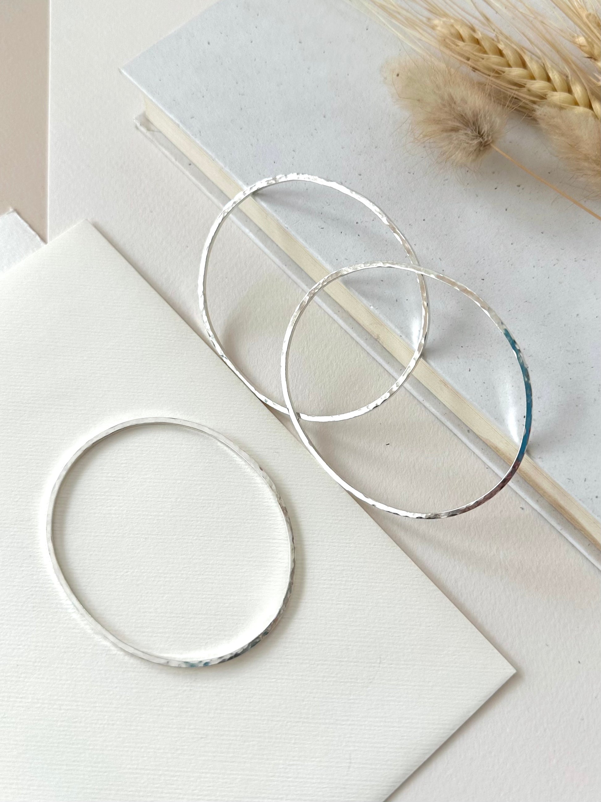 two sterling silvers bracelets leaning on the edge of a book and one displayed flat on a textured ivory background with dried grasses
