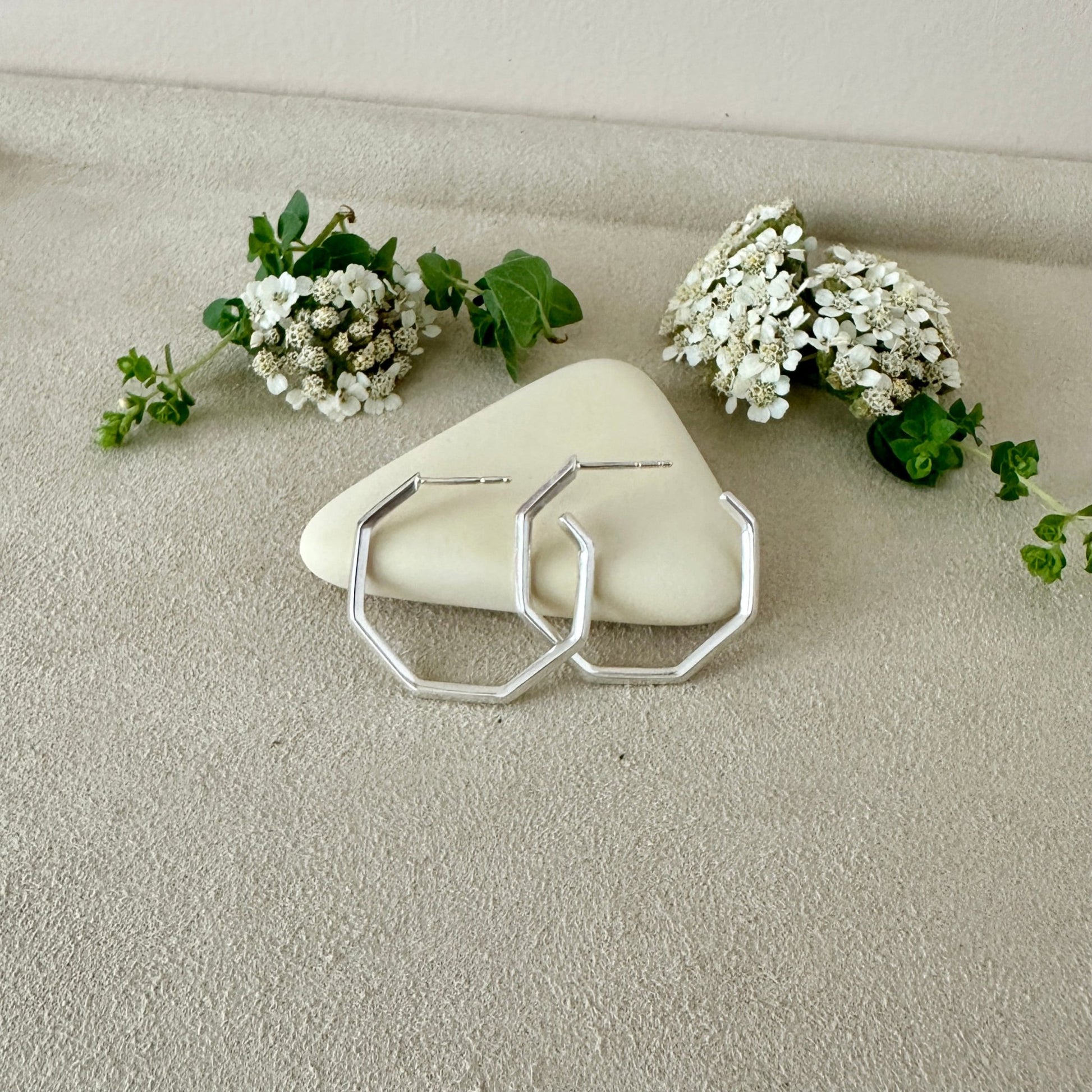 Silver geometric earrings on a stone with white flowers and green leaves.