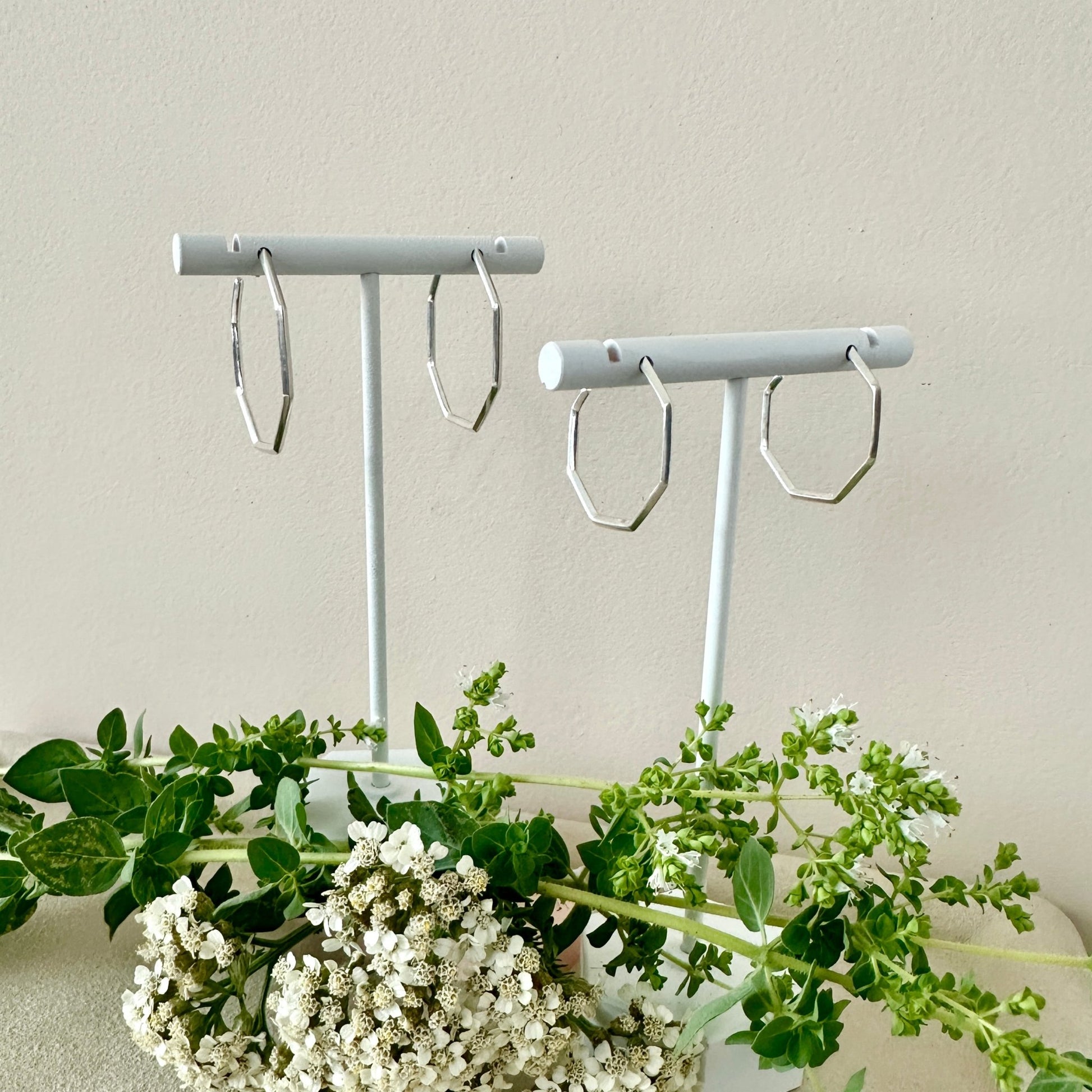 two pairs of hexagonal hoop earrings on white stands with a neutral background and greenery
