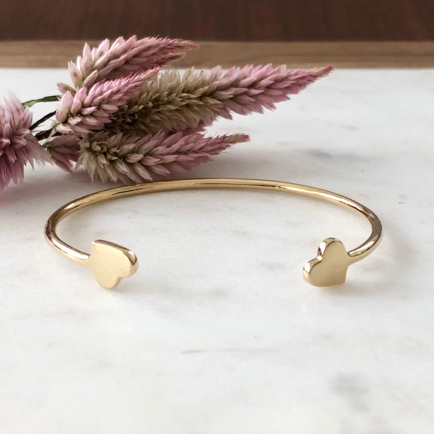 Gold cuff heart bracelet displayed on marble with a flower in the background.