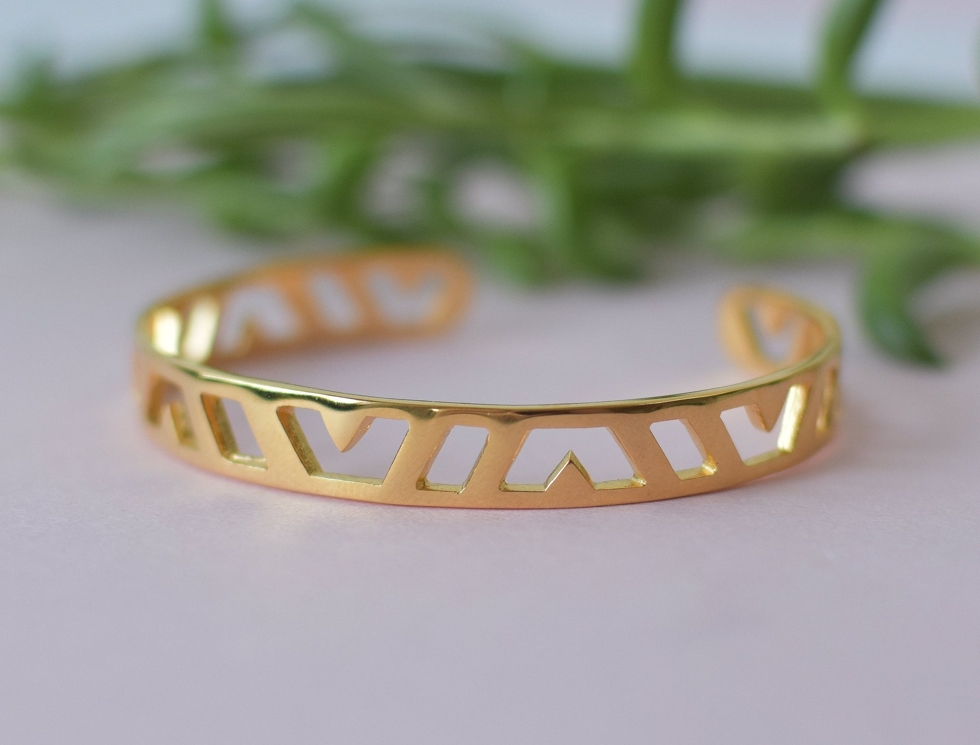 A close up shot off a gold cuff bracelet with geometric pattern and greenery in the background