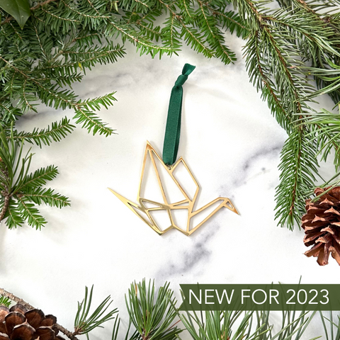 A handmade brass origami crane ornament on a marble background surrounded by greenery and pinecones