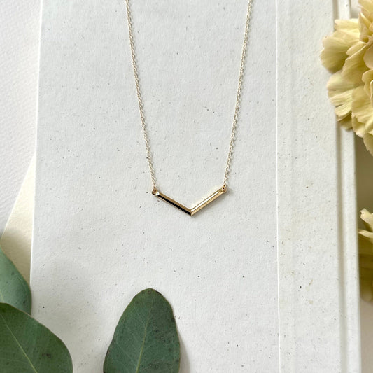 A handmade geometric v bar necklace in gold on white background with floral elements 