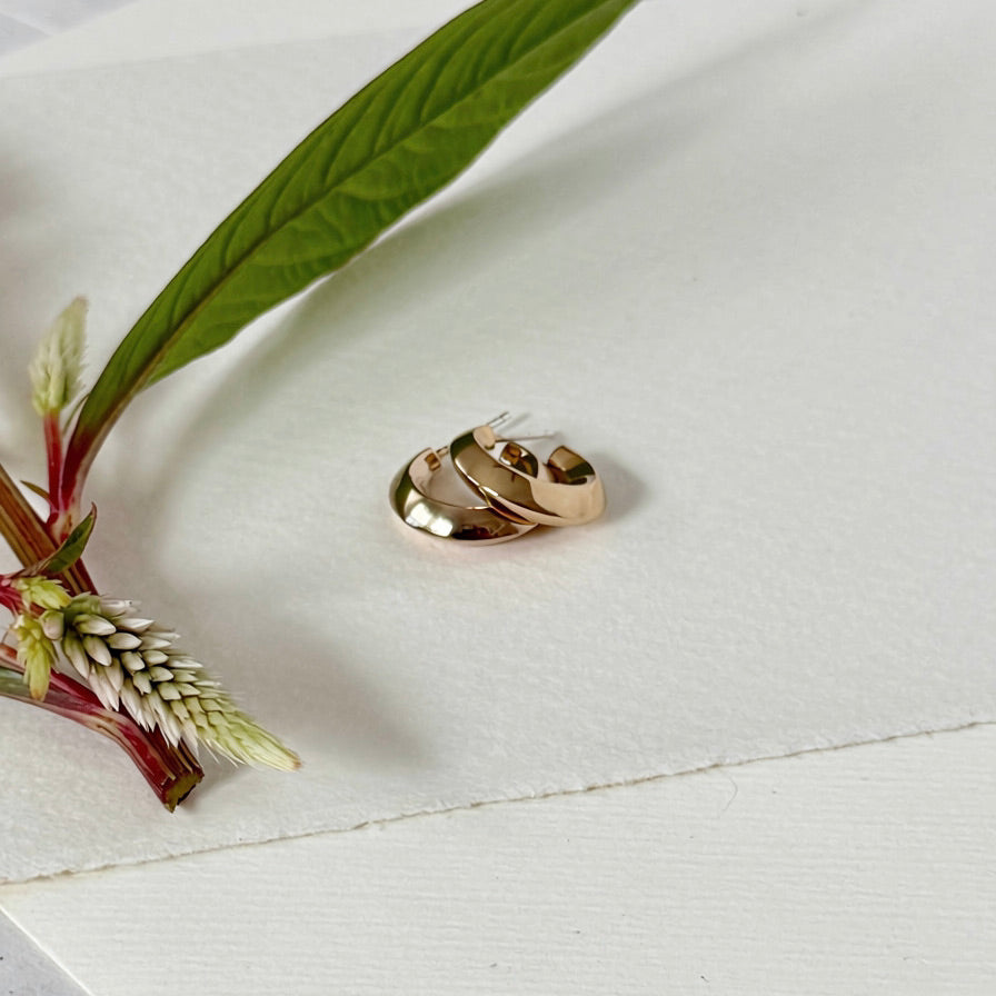 Two polished gold hoops stacked together on a textured white surface, styled with a green leaf and red flower stem.