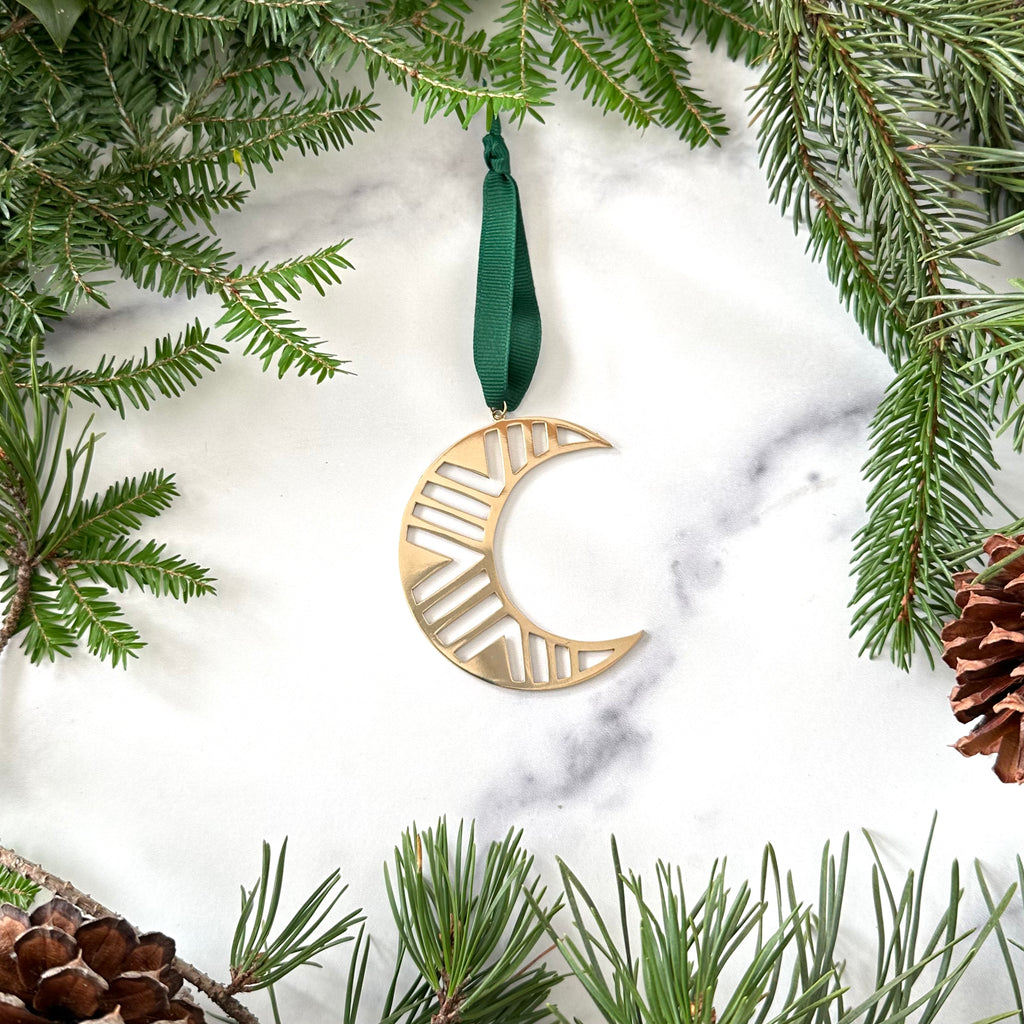 A handmade brass moon christmas ornament on a marble background surrounded by greenery with pinecones