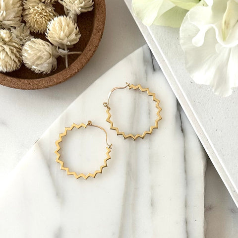 A pair of gold hoop earrings with a zig zag edge  displayed on a piece of marble with textured white background