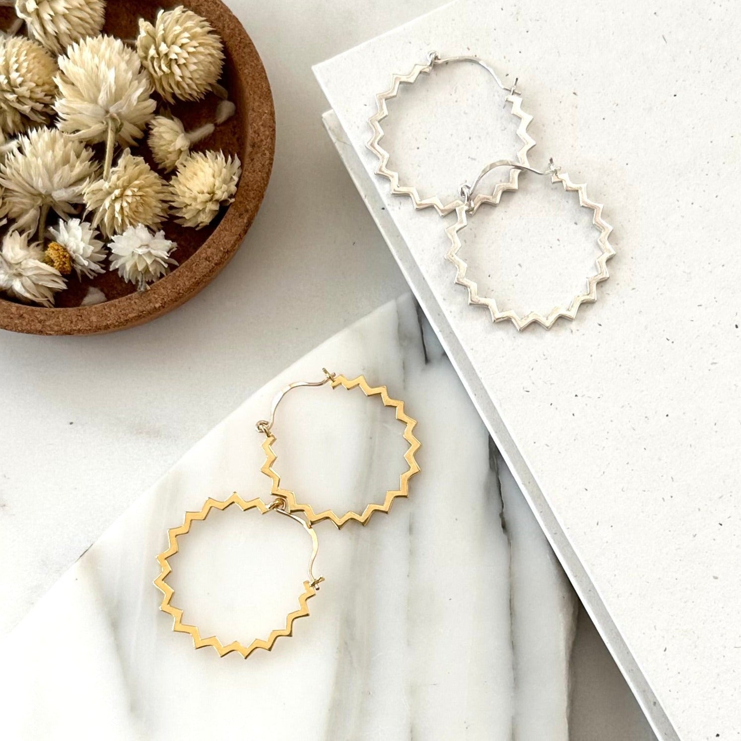 A pair of gold hoop and silver hoop mearrings with a zig zag edge displayed on a piece of marble with textured white background