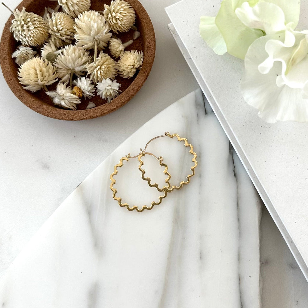 A pair of gold hoop earrings with a scallop edge displayed on a piece of marble with textured white background
