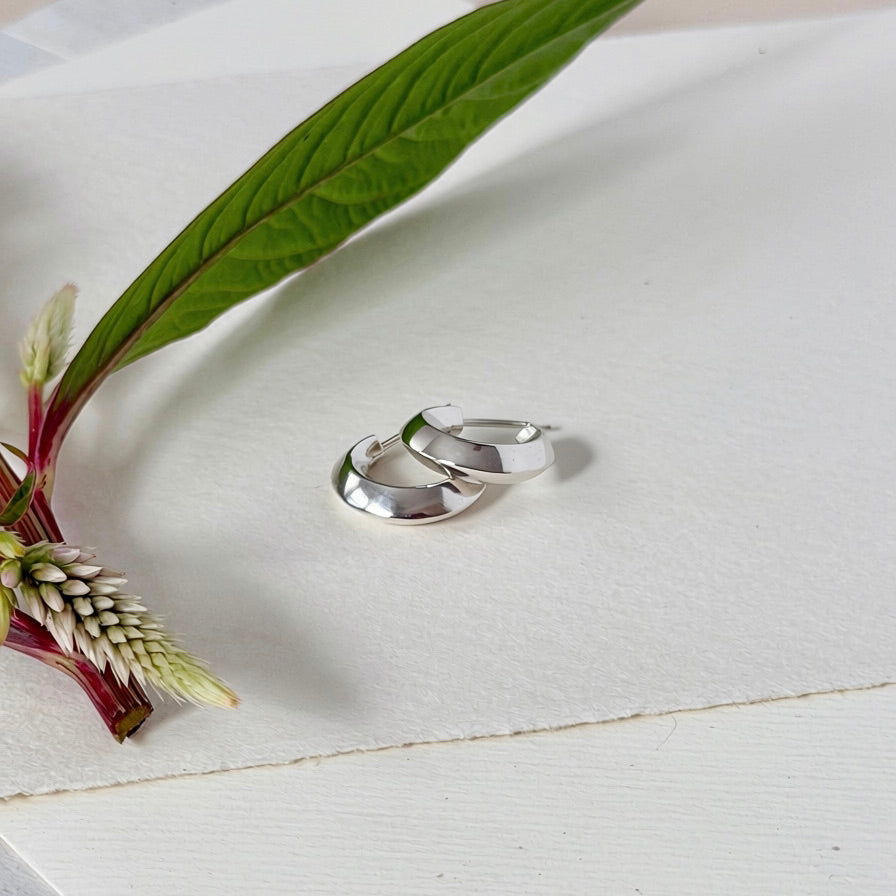 Two polished silver hoop earrings resting on a textured white surface, styled with a green leaf and a small white flower.