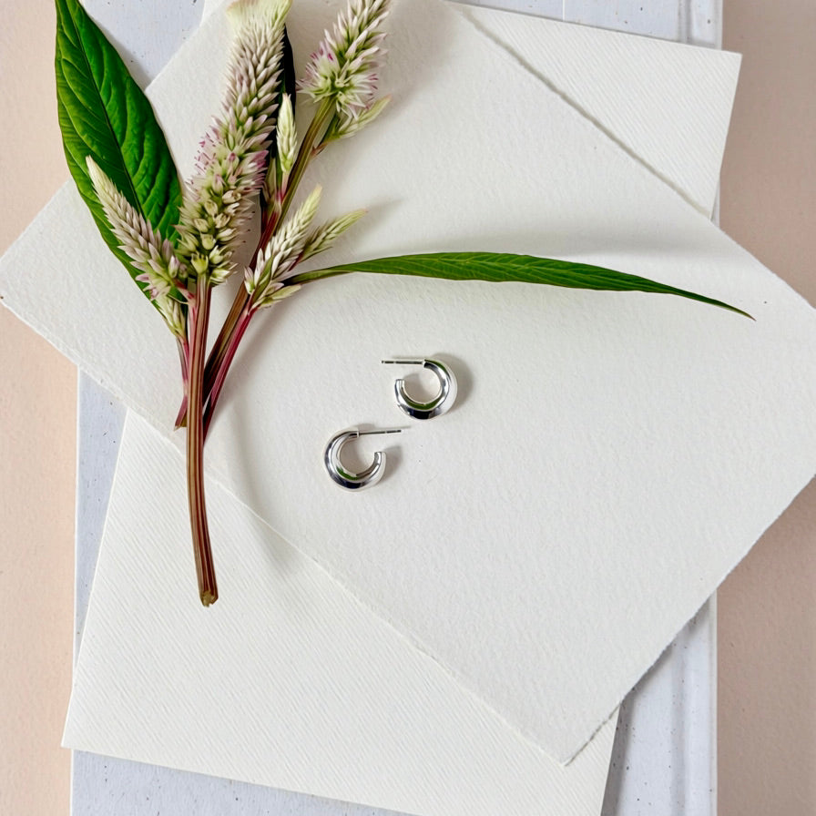 A pair of polished silver crescent hoop earrings displayed on a textured white paper envelope, styled with green leaves and pinkish-purple flowers.
