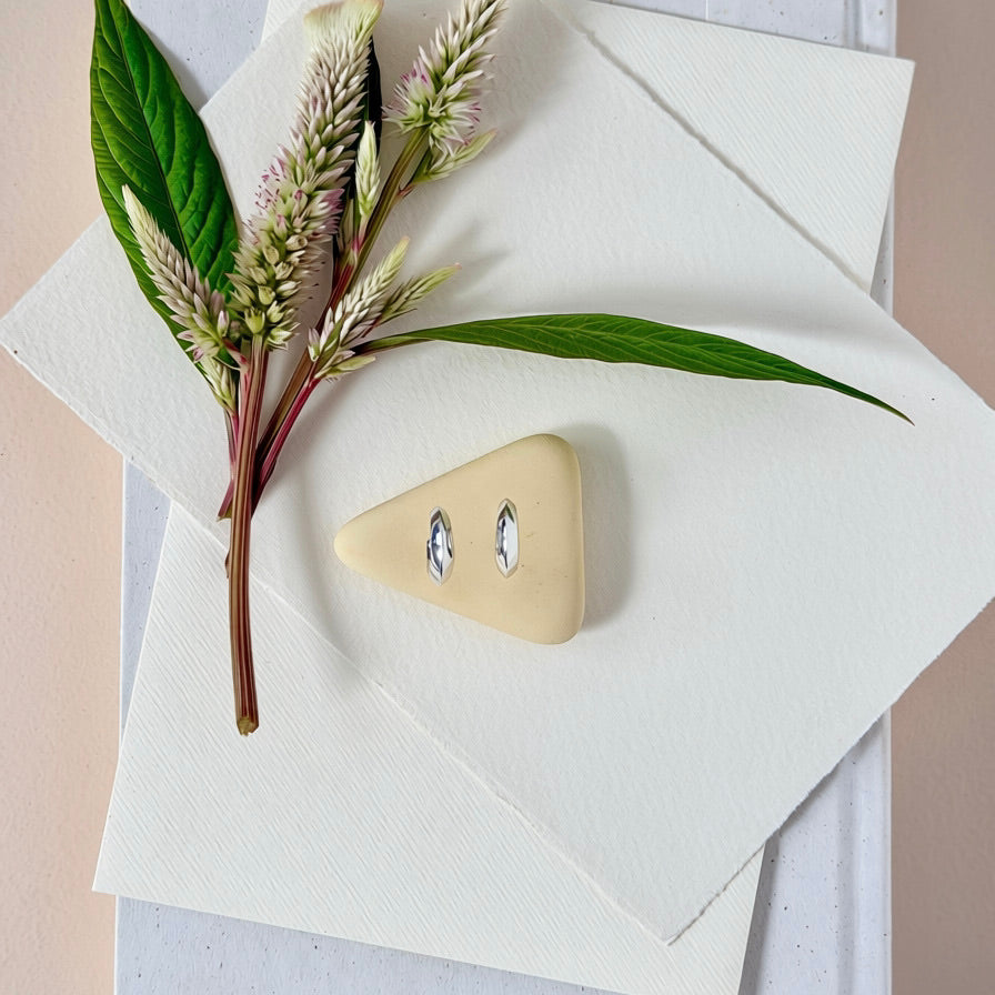 A pair of silver crescent shaped hoop earrings displayed on a small triangular cream stone, arranged on layered white textured paper with green leaves and dried flowers as styling props.