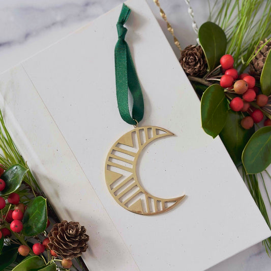 Gold crescent moon ornament with green ribbon on a white card with holly and pine cone decorations.