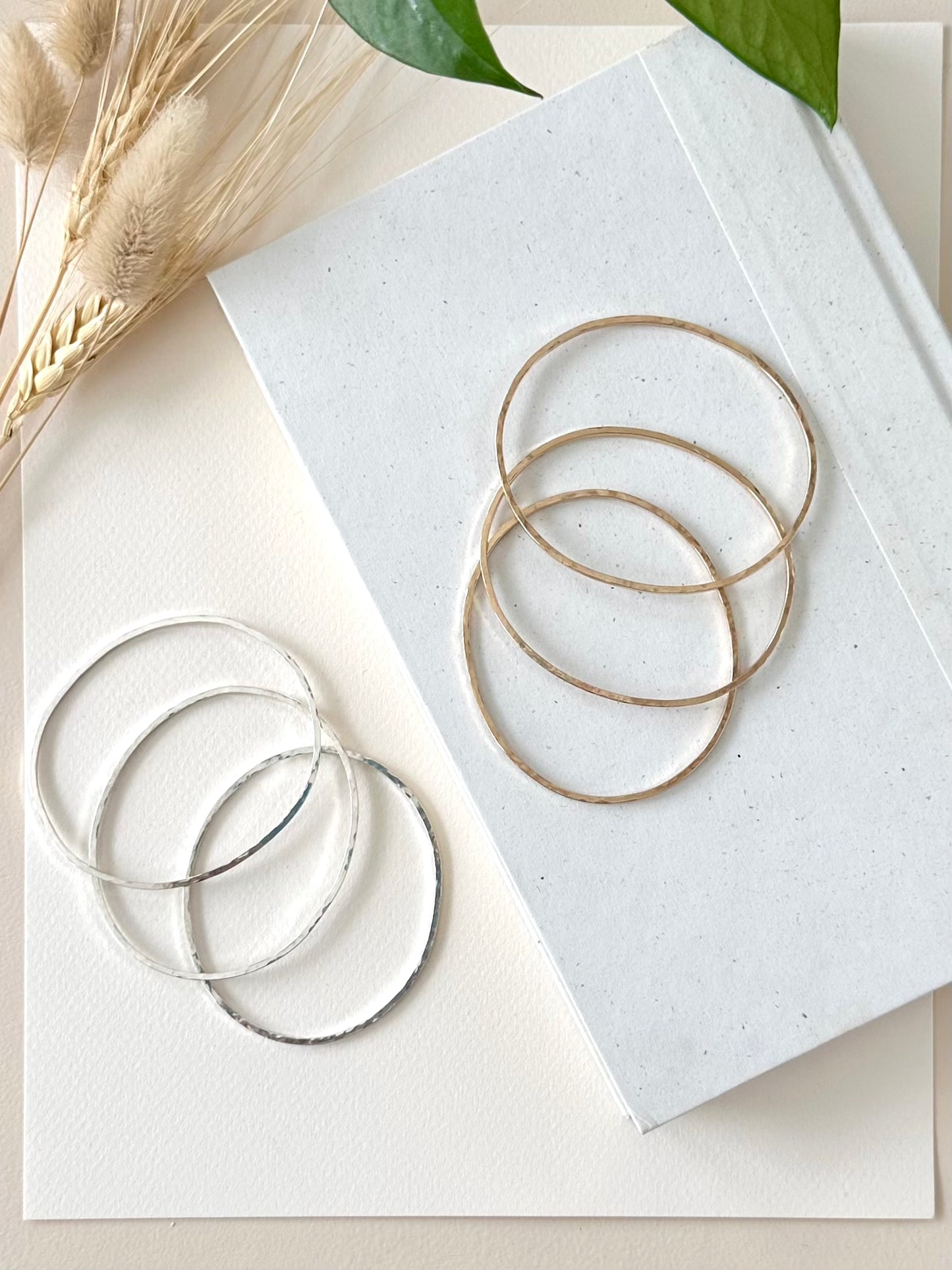 three gold bangle bracelets and three sterling silver bangle bracelets displayed on a textured white background with dried grasses and greenery