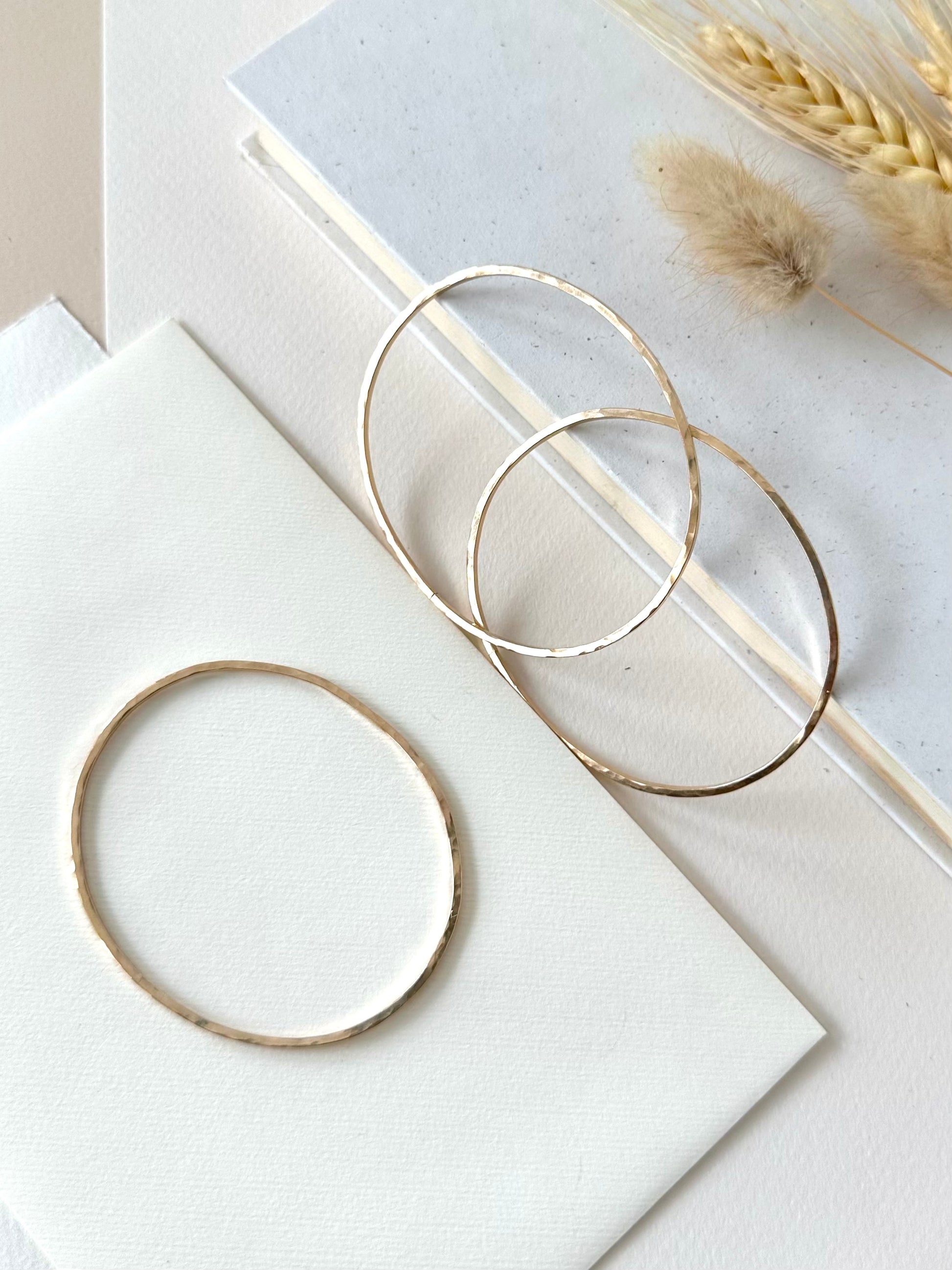 handmade hammered bangle bracelets, two displayed  leaning on a book and one laying flat on an ivory background with dried grasses 