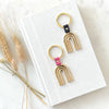 two brass key rings with arch design with leather detail in pink and black. the key rings are are on a neutral background with dried flowers