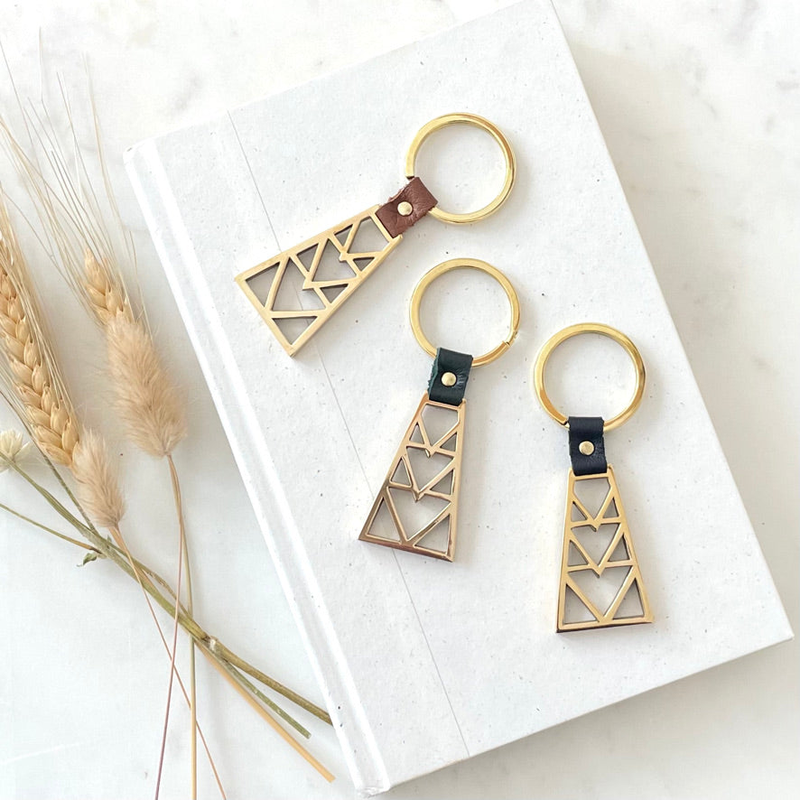 three brass key rings with a geometric design with leather detail in navy blue, brown, and  hunter green. the key rings are are on a neutral background with dried flowers
