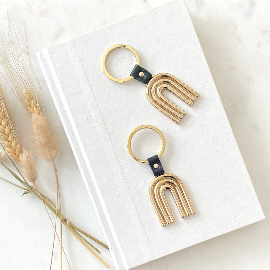 two brass key rings with arch design with leather detail in navy blue and hunter green. the key rings are are on a neutral background with dried flowers