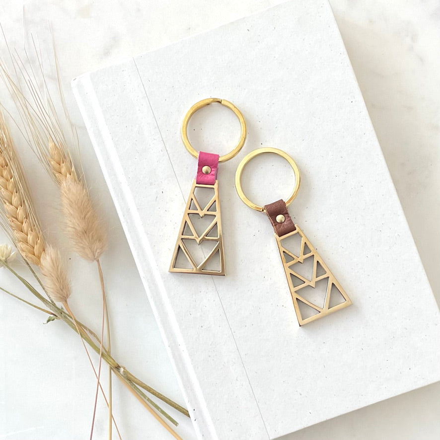 two brass key rings with a geometric design with leather detail in pink and brown. the key rings are are on a neutral background with dried flowers