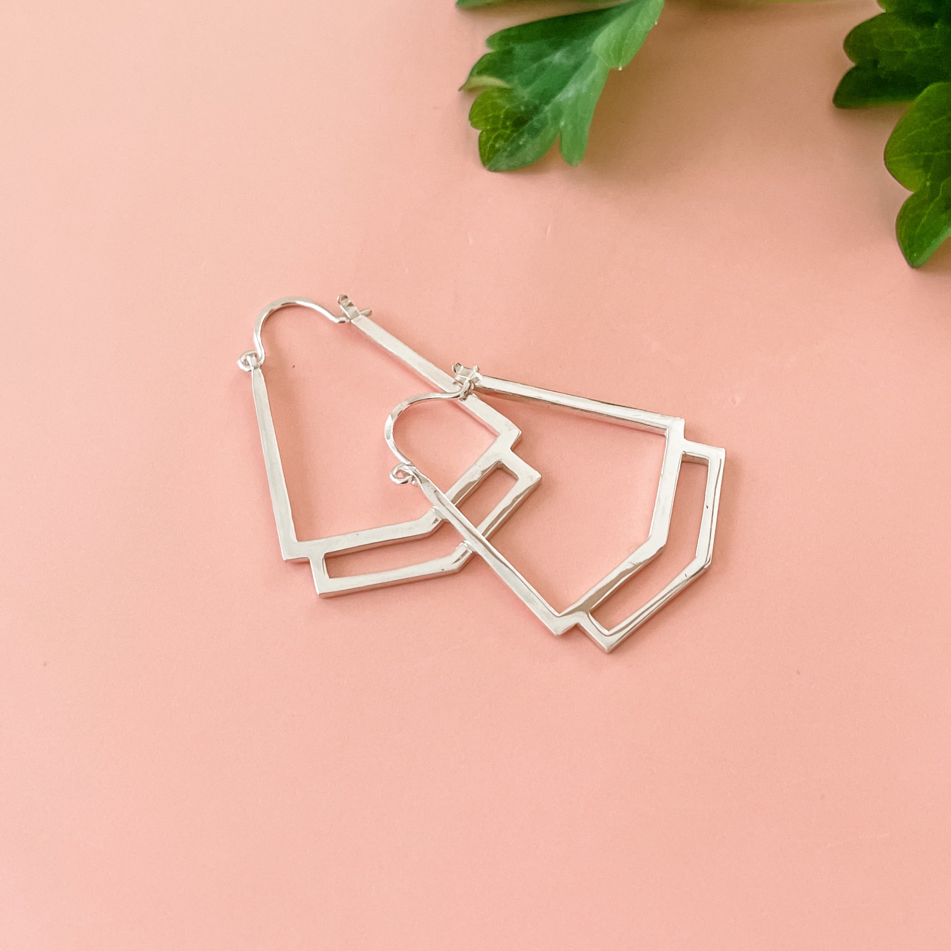 A close up of a pair of handmade sterling silver geometric hoop earrings displayed on a  pink background with greenery