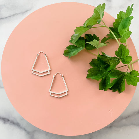 A pair of handmade sterling silver geometric hoop earrings displayed on a circular pink background with greenery