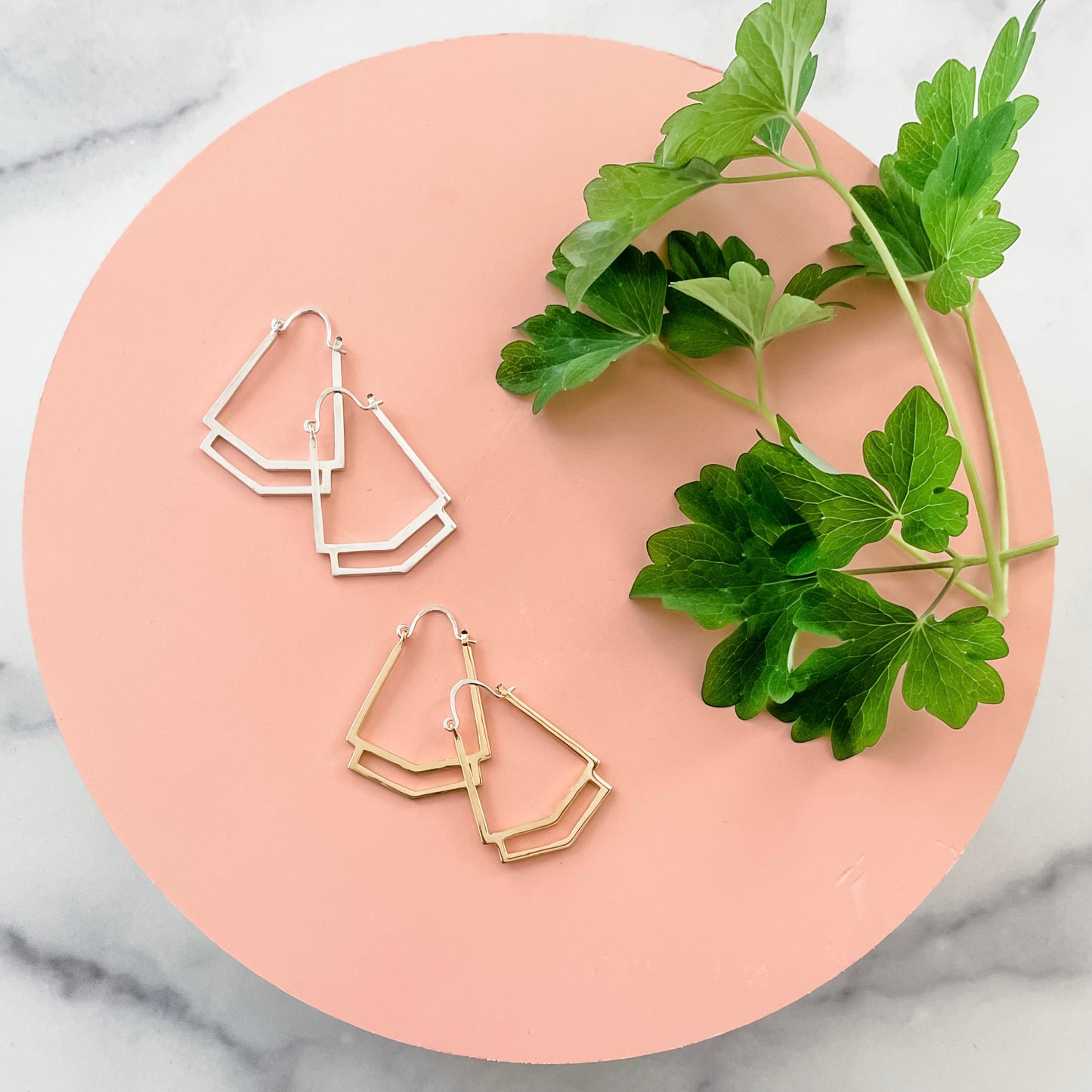 A pair of handmade sterling silver and gold geometric hoop earrings displayed on a circular pink background with greenery