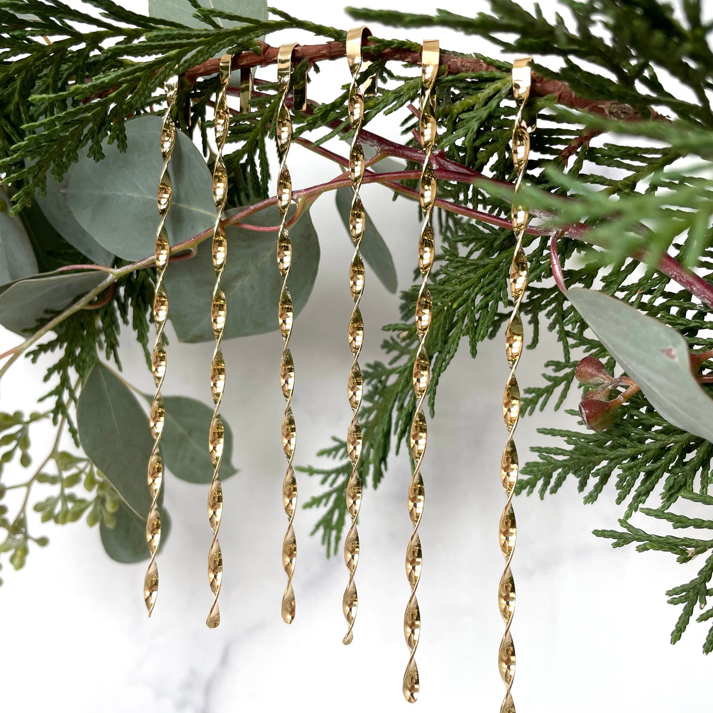 six gold plated christmas tree ornaments displayed on greenery with a white background