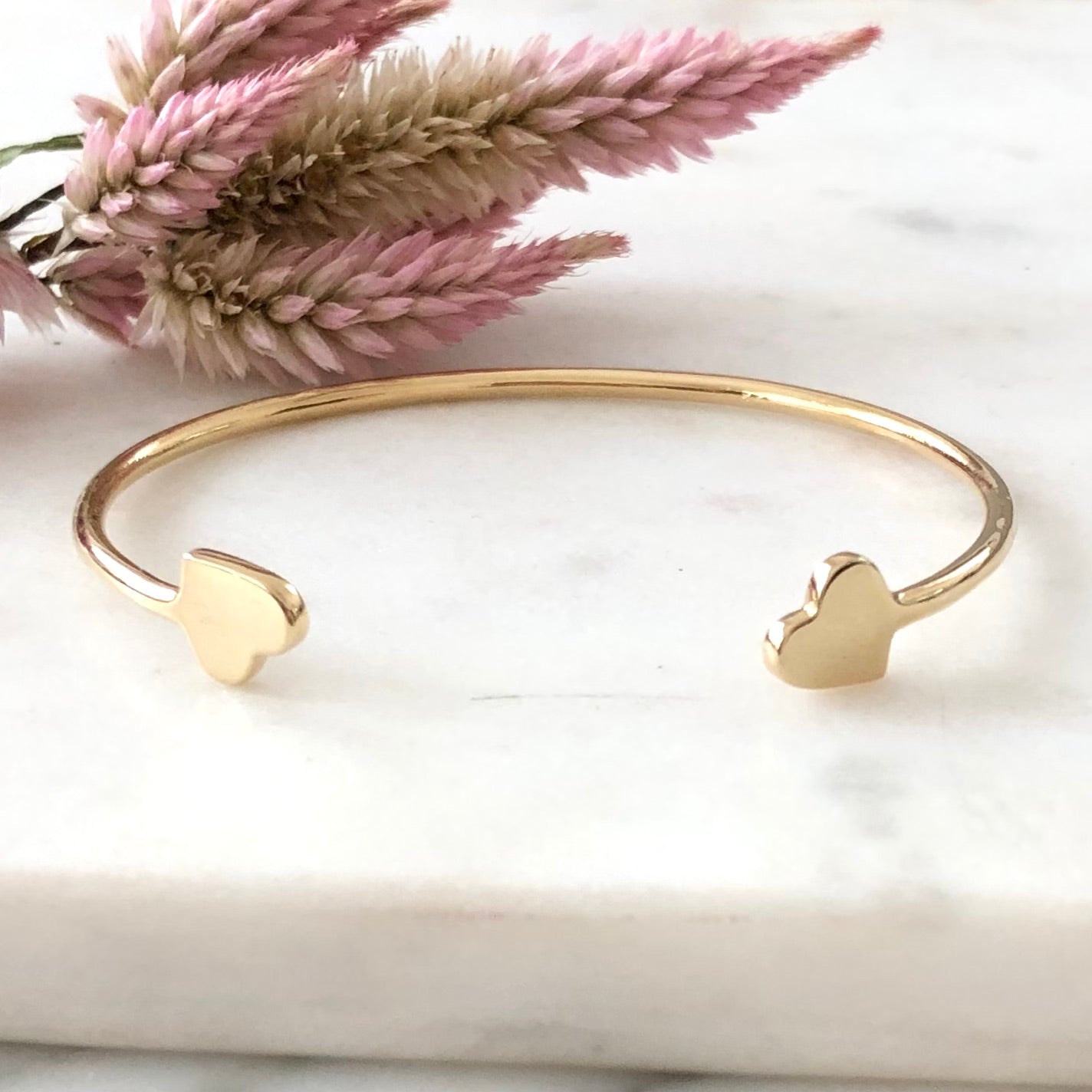 Gold cuff heart bracelet displayed on marble with a flower in the background.