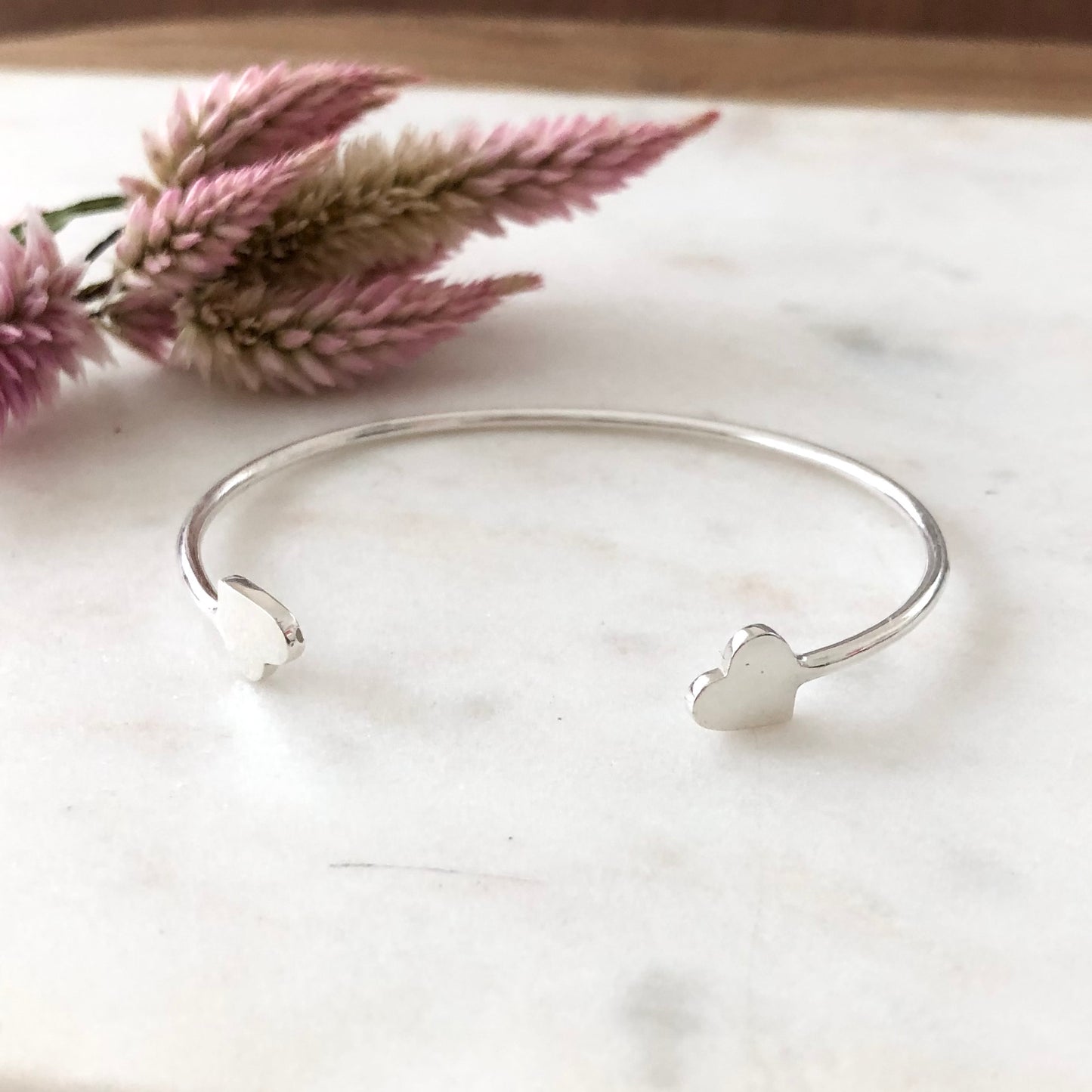 Silver cuff heart bracelet displayed on marble with a flower in the background.