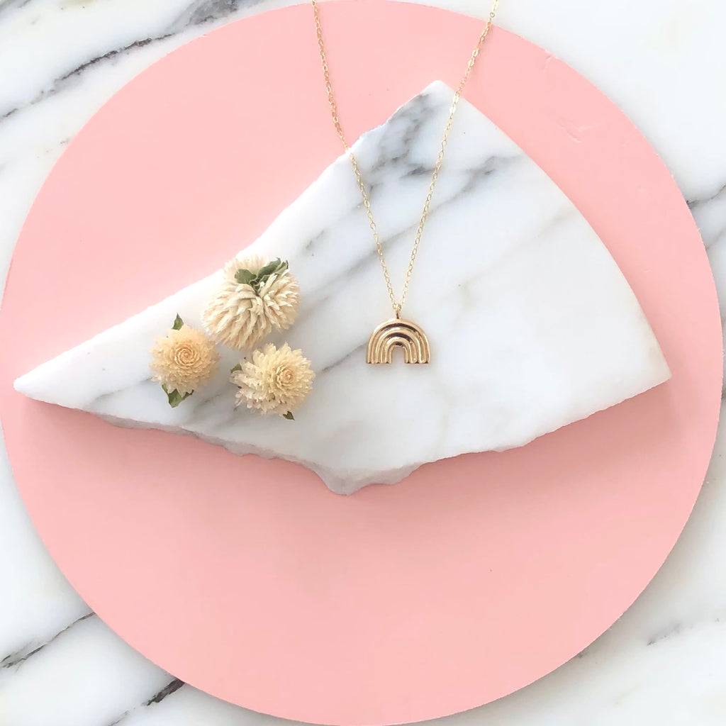 Gold rainbow necklace displayed on a piece of marble with pink circle background