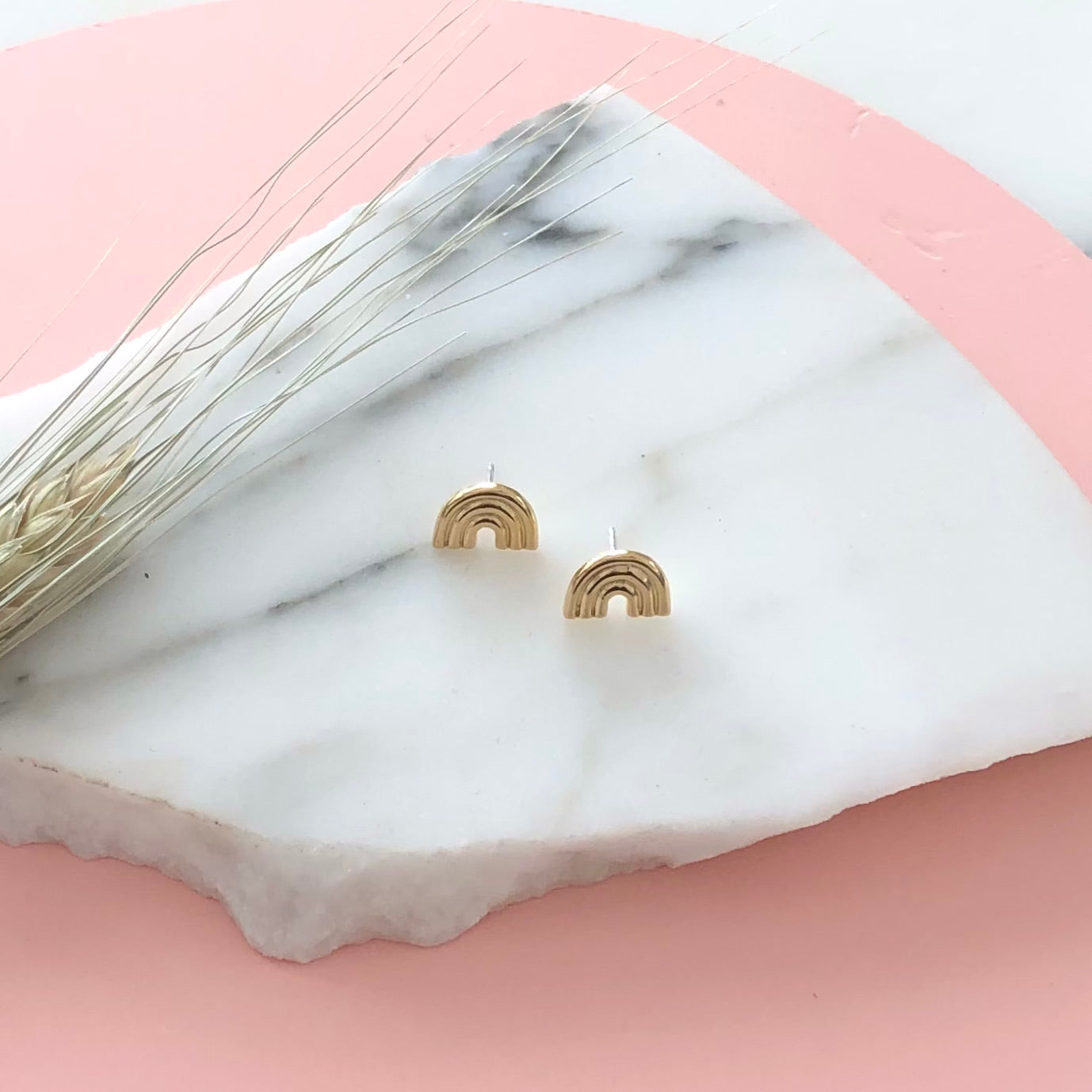 a pair of gold rainbow stud earrings displayed on a piece of marble with a pink background