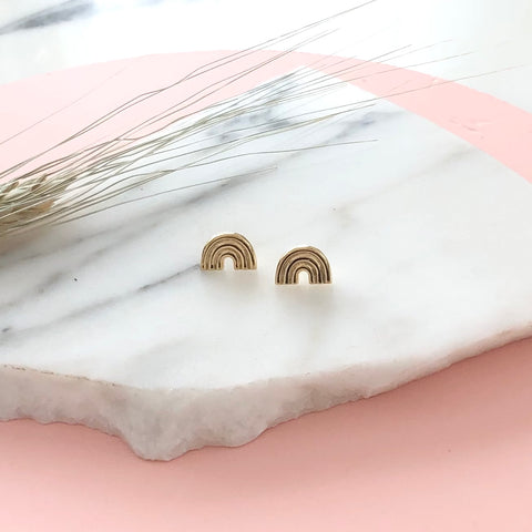 a pair of gold rainbow stud earrings displayed on a piece of marble with a pink background 