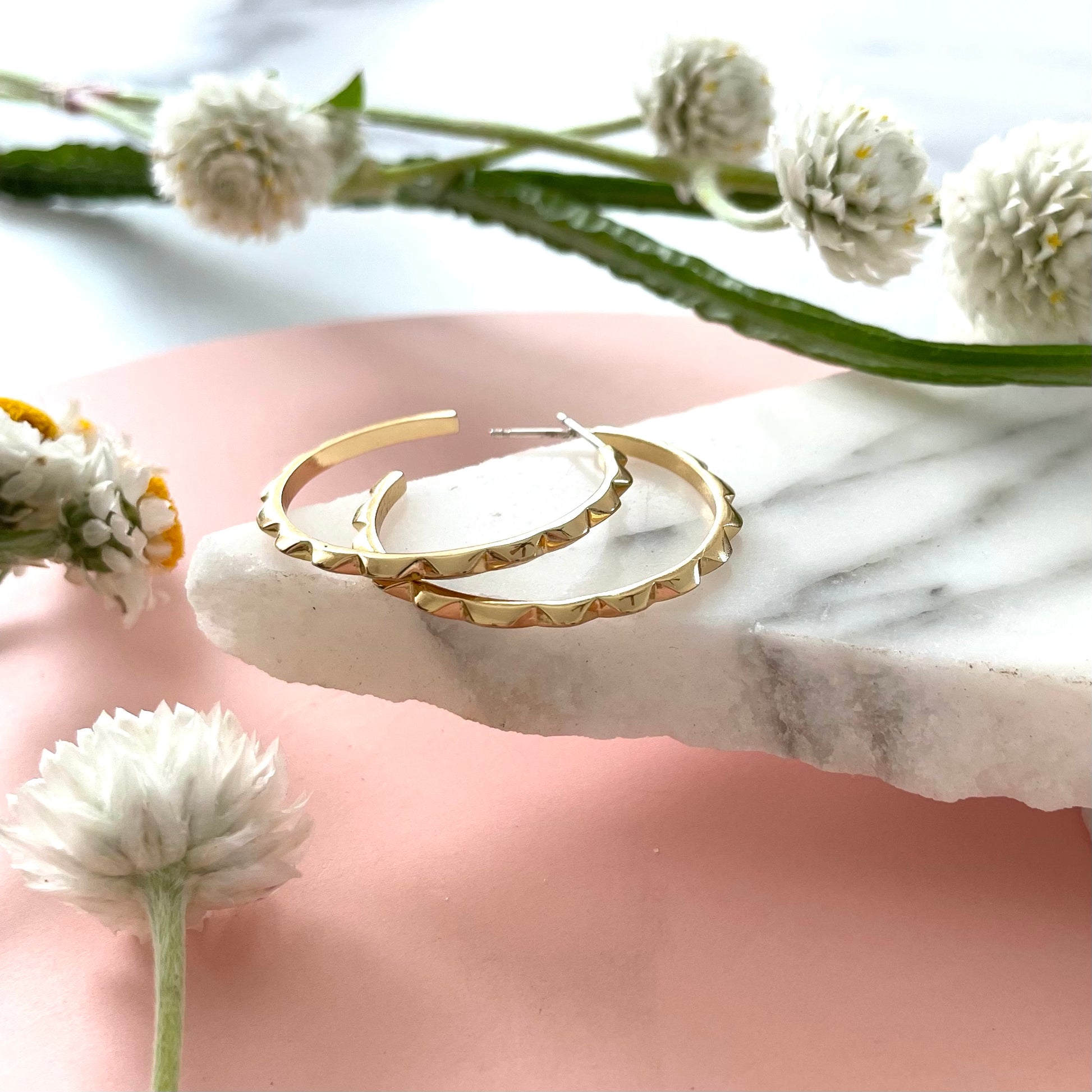 Handmade gold hoop earrings on a marble displayed with a pink background and fresh flowers