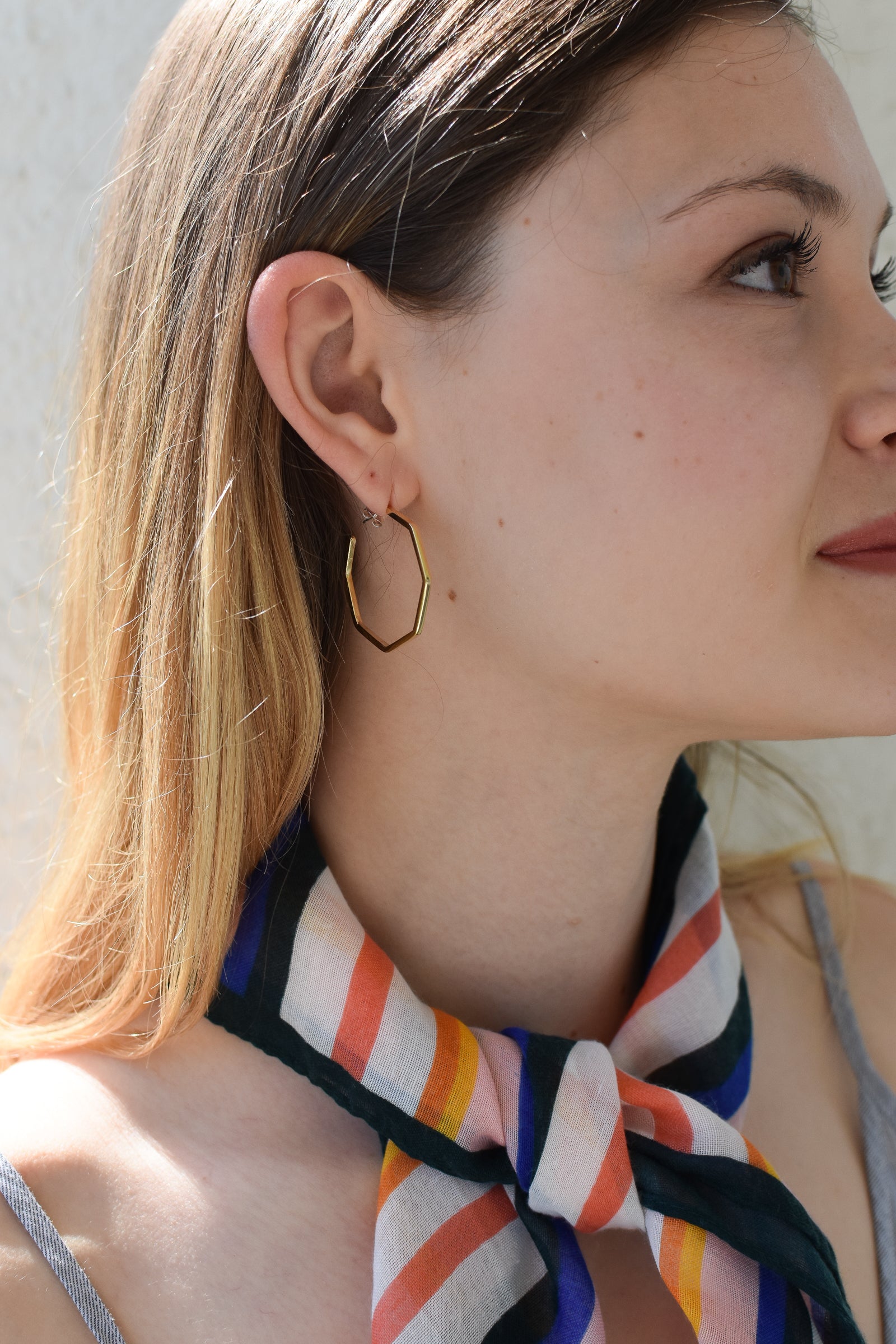 A pair geometric gold hoop earrings on a female model with blonde hair wearing a multi stripe scarf