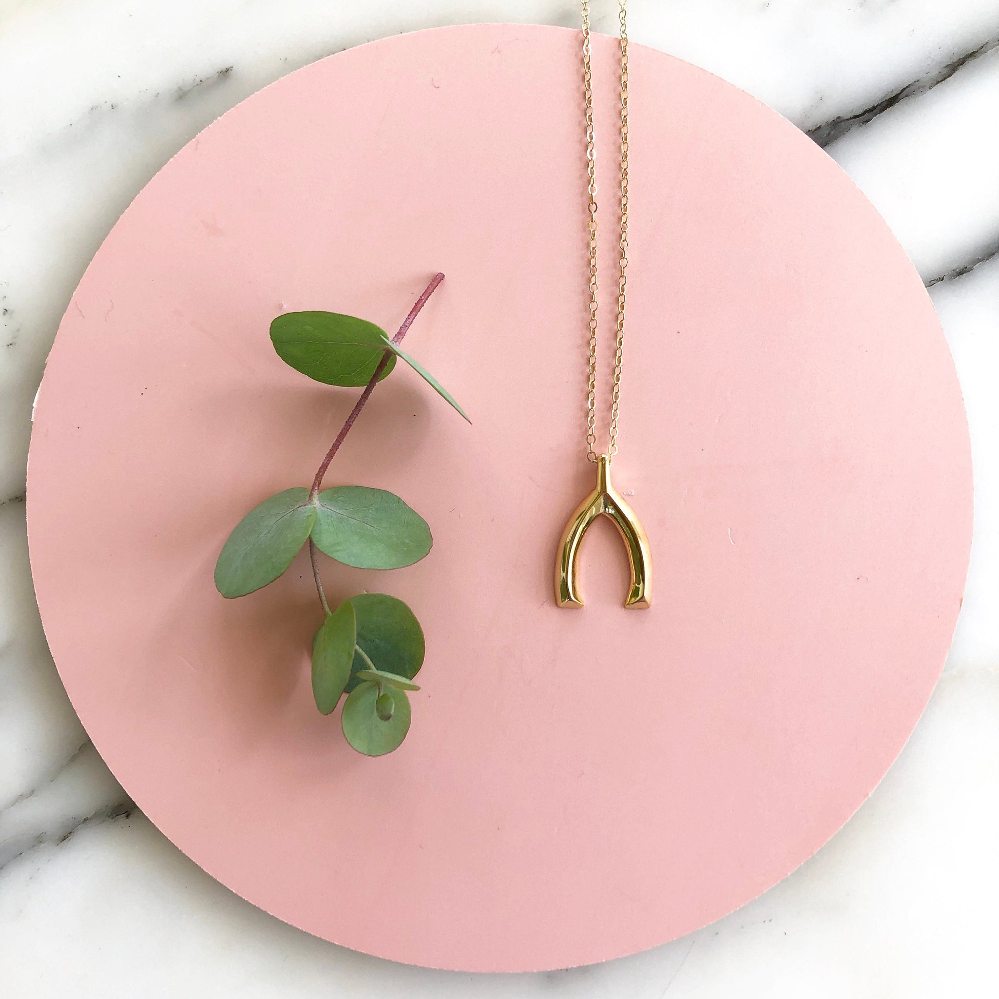 Sarah Cecelia Gold wishbone necklace  on a circular pink background with marble and eucalyptus