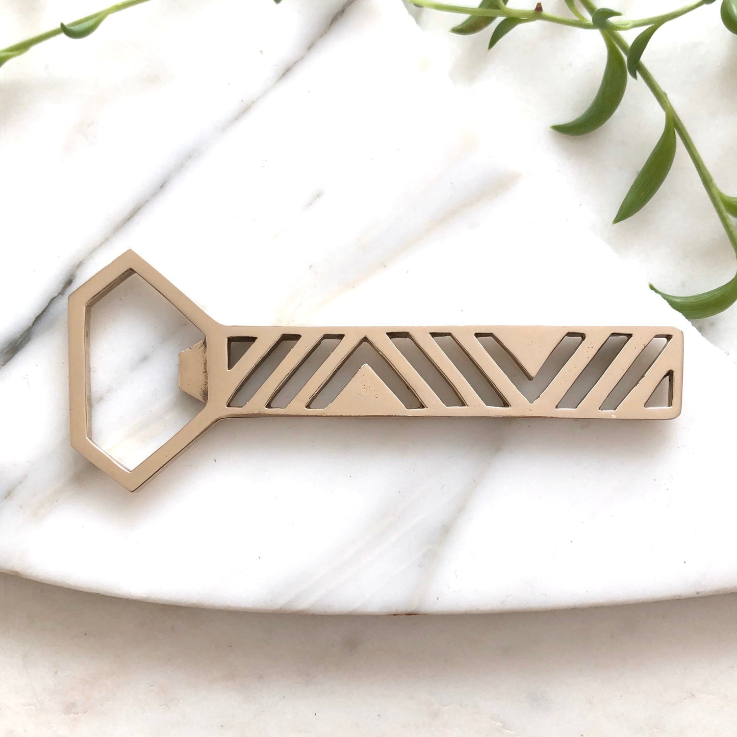A handmade brass bottle opener by Sarah Cecelia on a marble slab with greenery