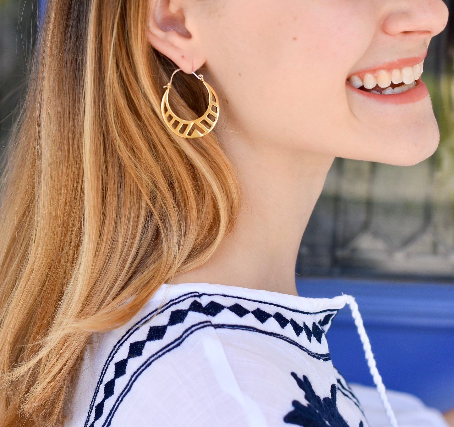 Gold hoop earrings with a cut geometric pattern on a model with blonde hair and a blue and white top