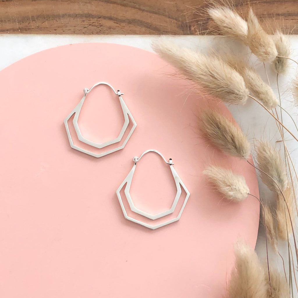 A pair of handmade geometric sterling silver hoop earrings on a pink background with dried flowers 