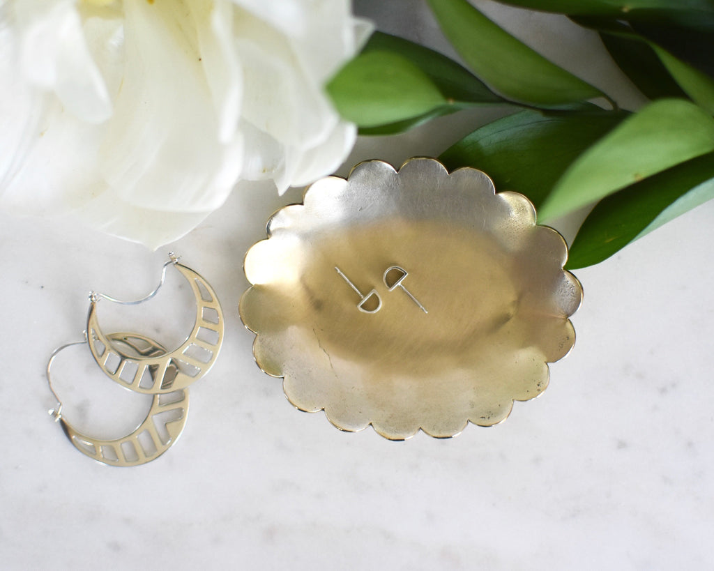 A scalloped edge brass jewelry dish holding stud earrings with silver hoops beside the dish and florals in the background 